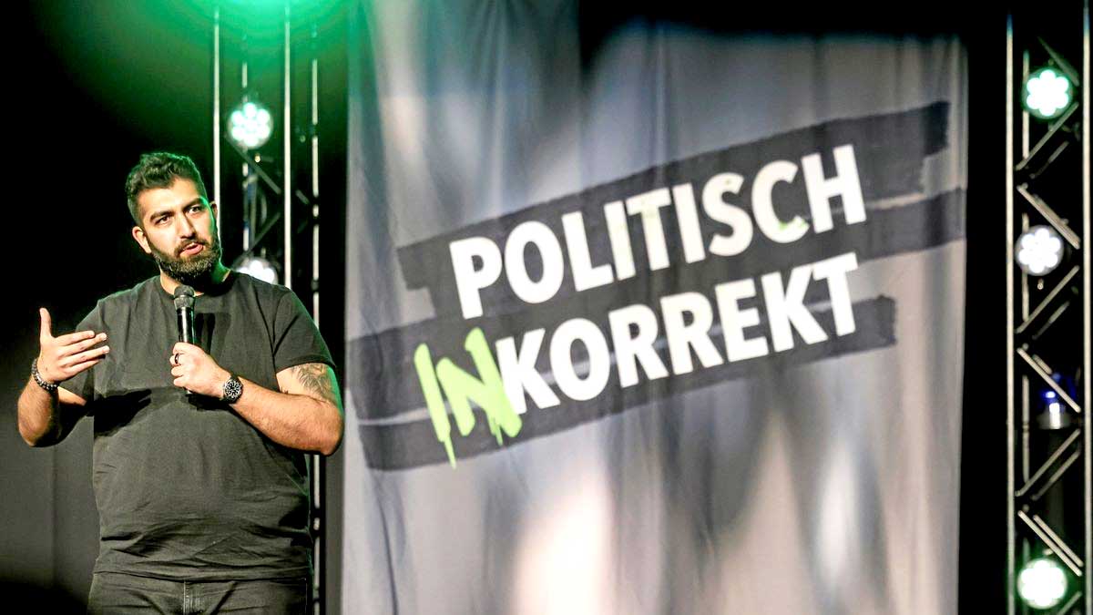 Faisal Kawusi auf der Bühne mit Politisch inkorrekt Banner im Hintergrund